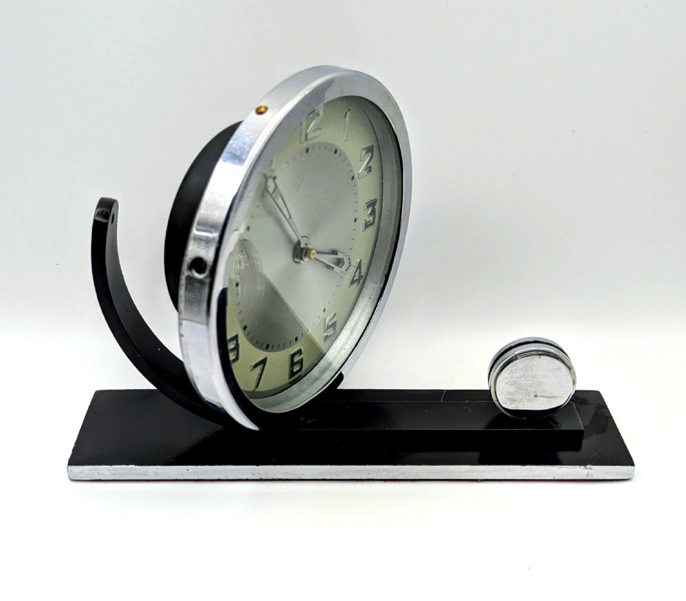 An Art Deco circular desk clock with a chrome bezel and silver dial mounted on an asymmetrical black rectangular base. View V