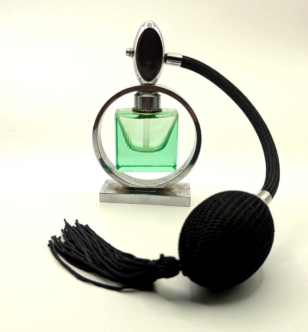 An Art Deco perfume atomiser featuring a square green glass bottle suspended inside a circular chrome frame on a rectangular base, connected to a black woven squeeze bulb with a tassel.