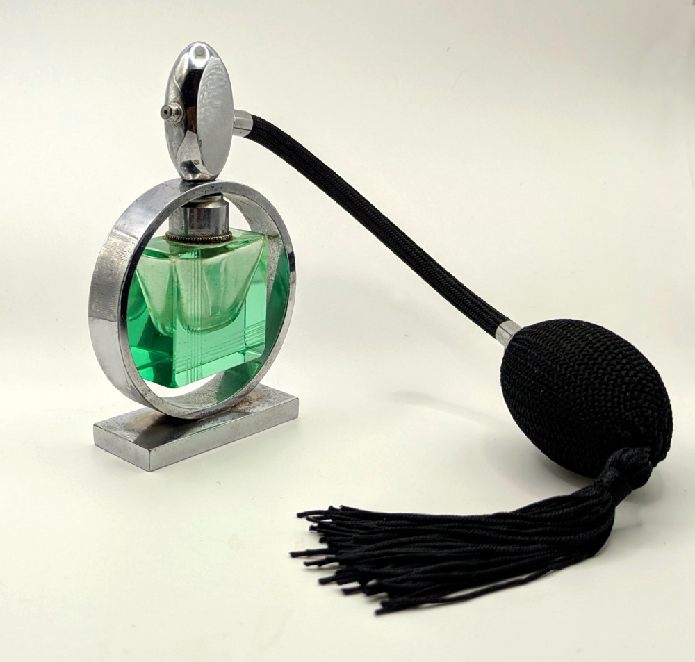 An Art Deco perfume atomiser featuring a square green glass bottle suspended inside a circular chrome frame on a rectangular base, connected to a black woven squeeze bulb with a tassel. View III