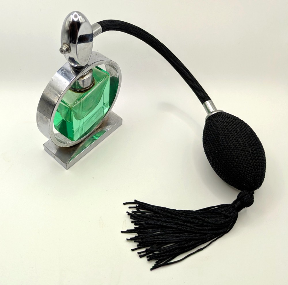 An Art Deco perfume atomiser featuring a square green glass bottle suspended inside a circular chrome frame on a rectangular base, connected to a black woven squeeze bulb with a tassel. View IV