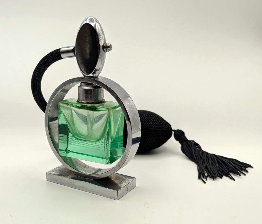 An Art Deco perfume atomiser featuring a square green glass bottle suspended inside a circular chrome frame on a rectangular base, connected to a black woven squeeze bulb with a tassel. View V