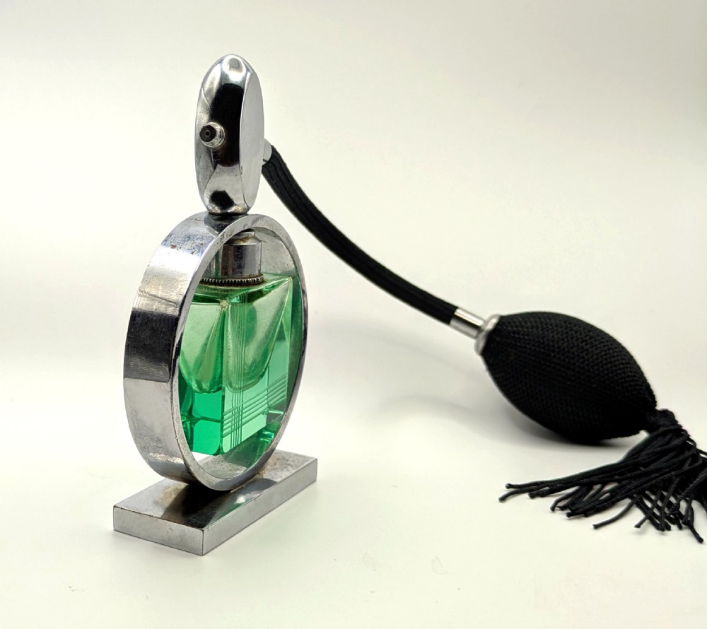 An Art Deco perfume atomiser featuring a square green glass bottle suspended inside a circular chrome frame on a rectangular base, connected to a black woven squeeze bulb with a tassel. View VI