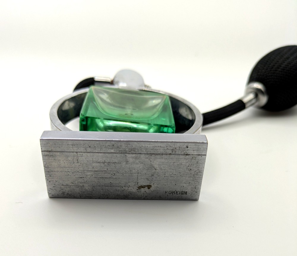 An Art Deco perfume atomiser featuring a square green glass bottle suspended inside a circular chrome frame on a rectangular base, connected to a black woven squeeze bulb with a tassel. View VIII