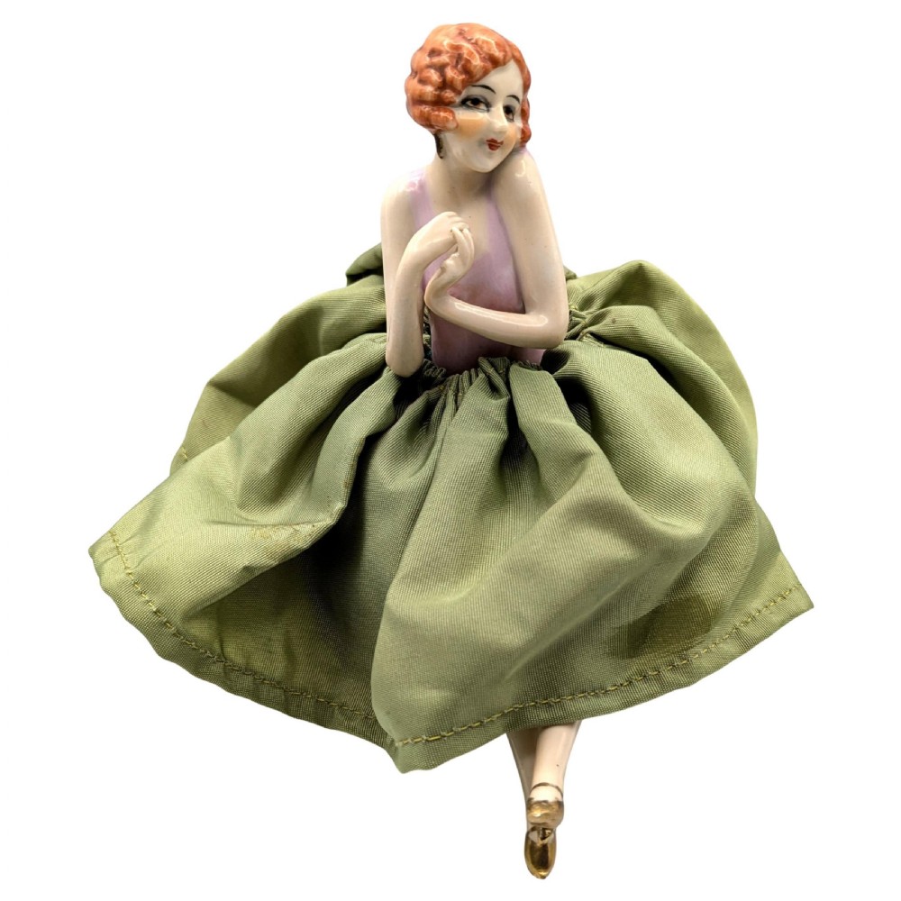 An Art Deco porcelain half doll depicting a flapper girl with auburn bobbed hair, a pink bodice, a voluminous sage green fabric skirt, and fine porcelain legs.