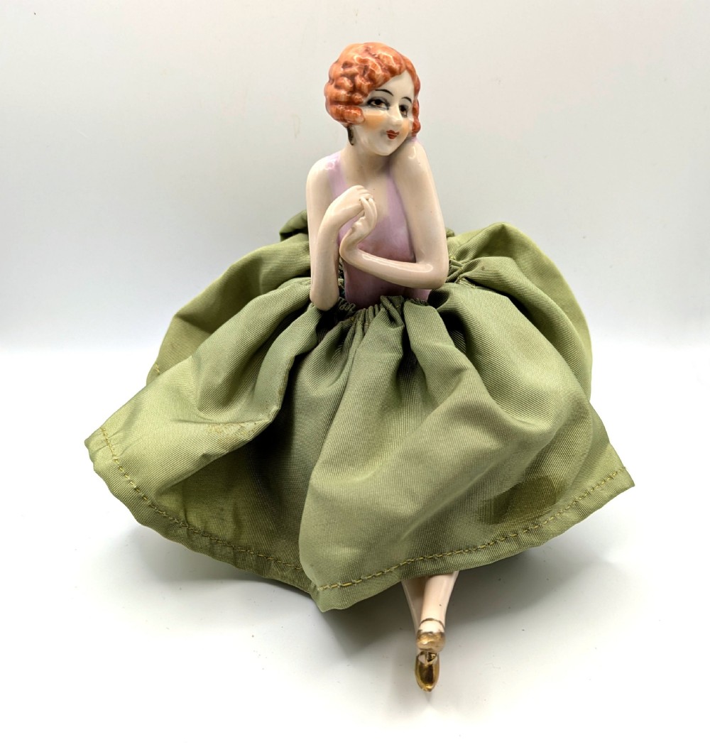 An Art Deco porcelain half doll depicting a flapper girl with auburn bobbed hair, a pink bodice, a voluminous sage green fabric skirt, and fine porcelain legs. View II