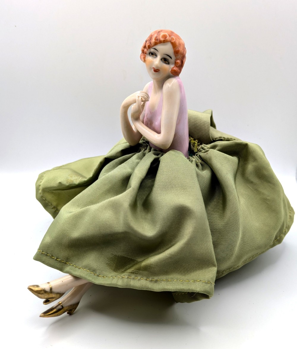 An Art Deco porcelain half doll depicting a flapper girl with auburn bobbed hair, a pink bodice, a voluminous sage green fabric skirt, and fine porcelain legs. View III