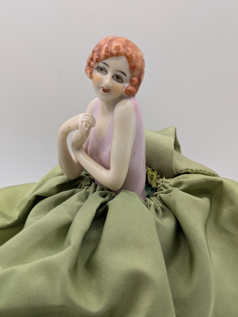An Art Deco porcelain half doll depicting a flapper girl with auburn bobbed hair, a pink bodice, a voluminous sage green fabric skirt, and fine porcelain legs. View IV