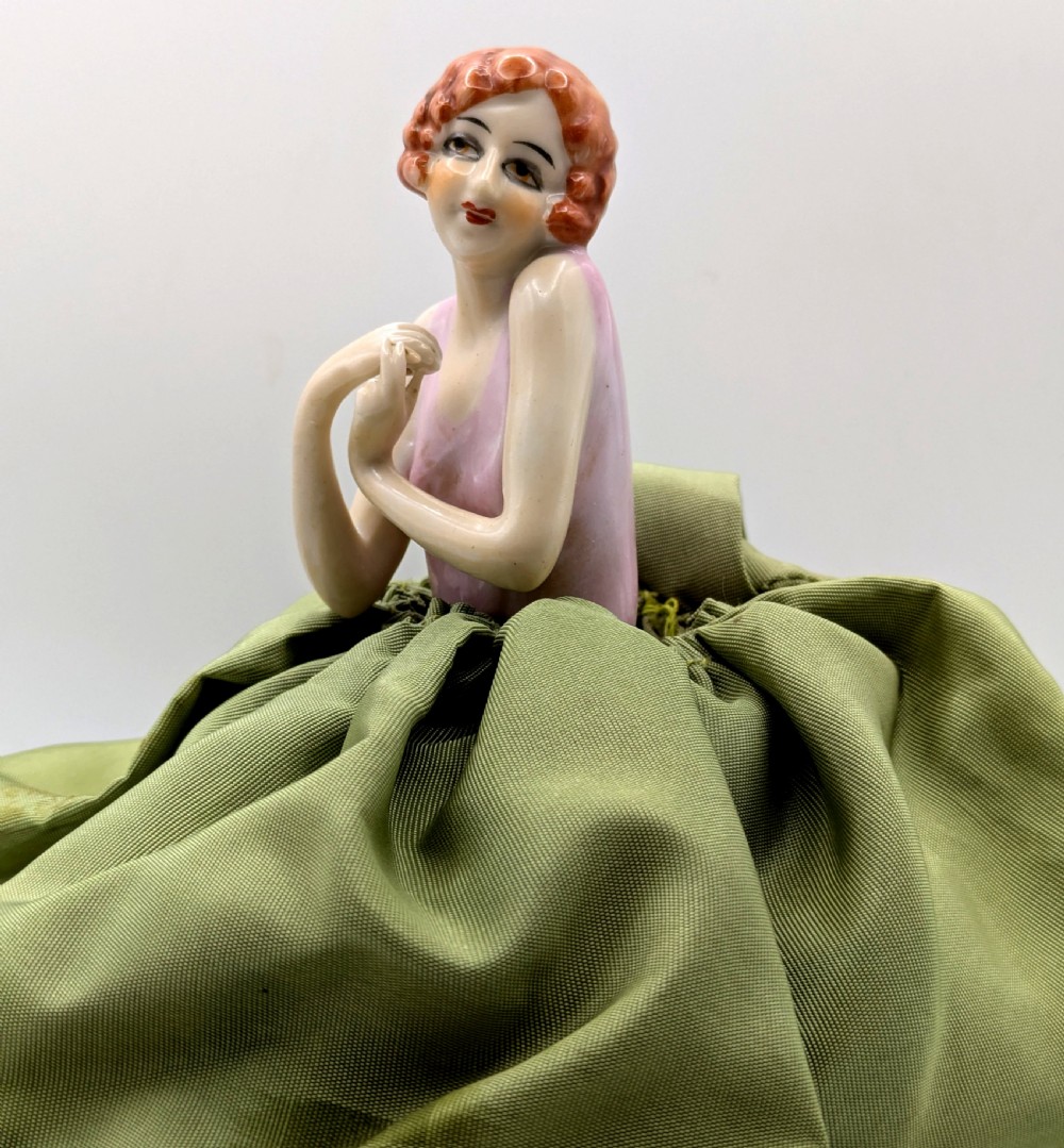 An Art Deco porcelain half doll depicting a flapper girl with auburn bobbed hair, a pink bodice, a voluminous sage green fabric skirt, and fine porcelain legs. View V