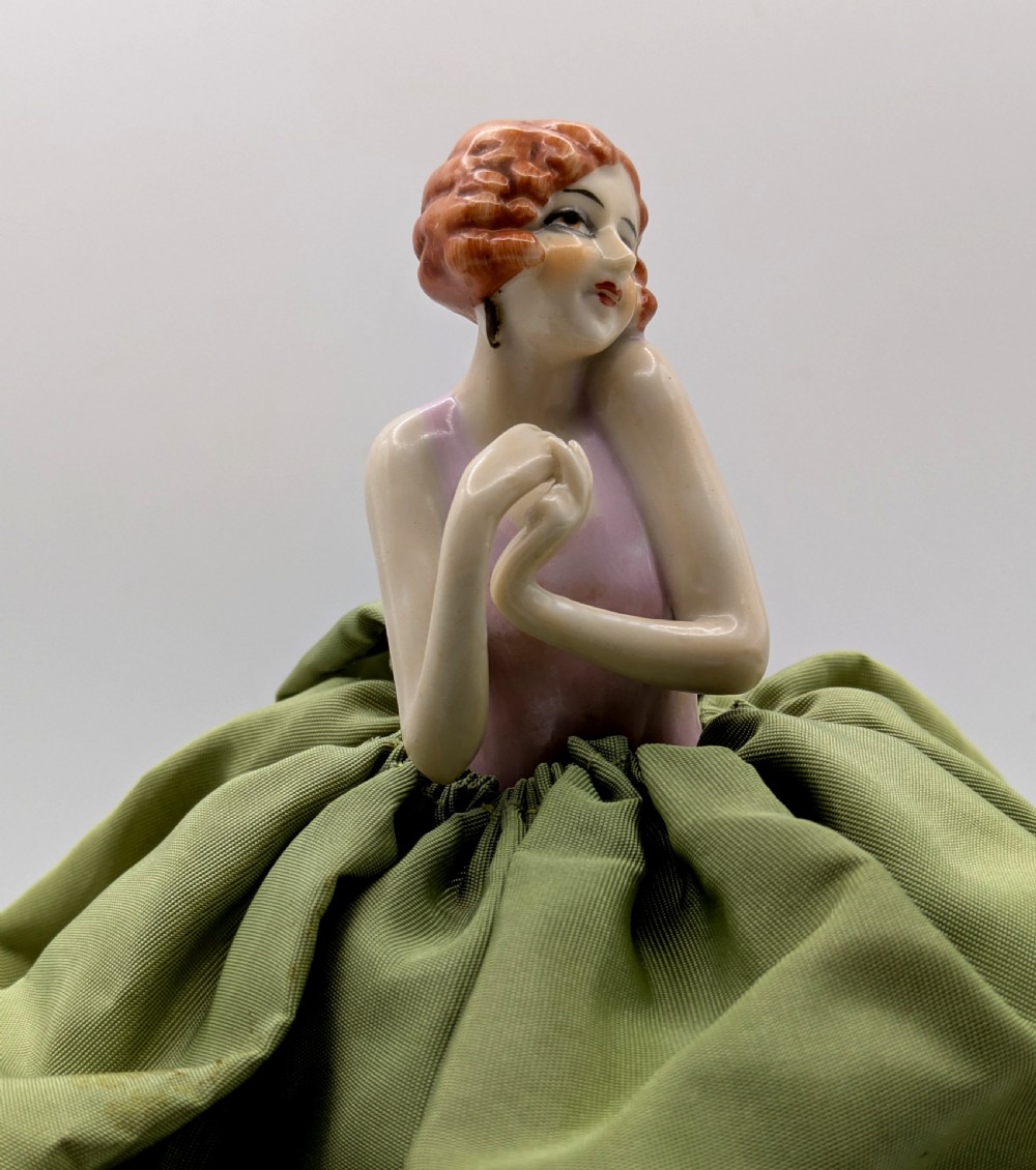 An Art Deco porcelain half doll depicting a flapper girl with auburn bobbed hair, a pink bodice, a voluminous sage green fabric skirt, and fine porcelain legs. View VI