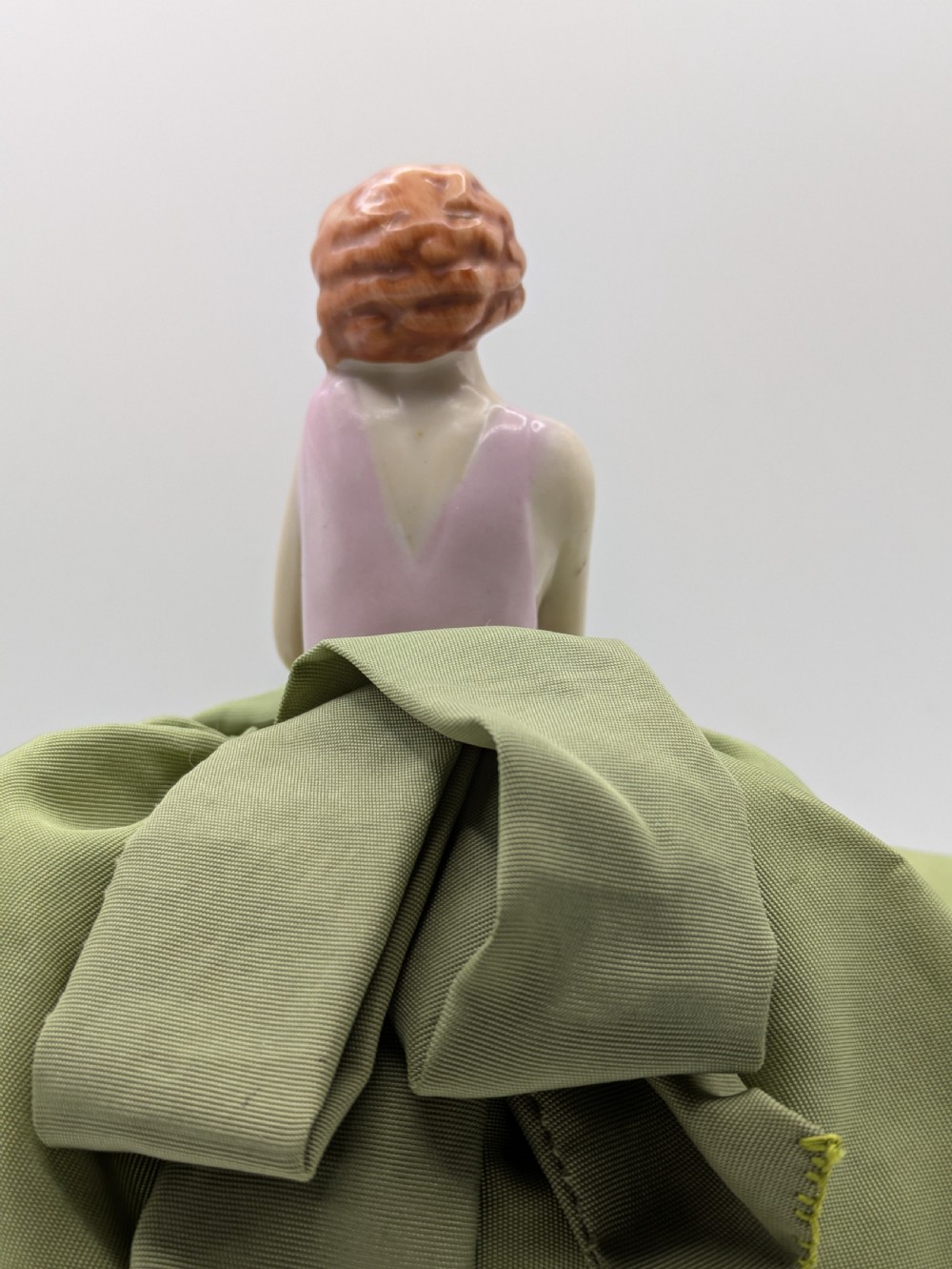 An Art Deco porcelain half doll depicting a flapper girl with auburn bobbed hair, a pink bodice, a voluminous sage green fabric skirt, and fine porcelain legs. View VII