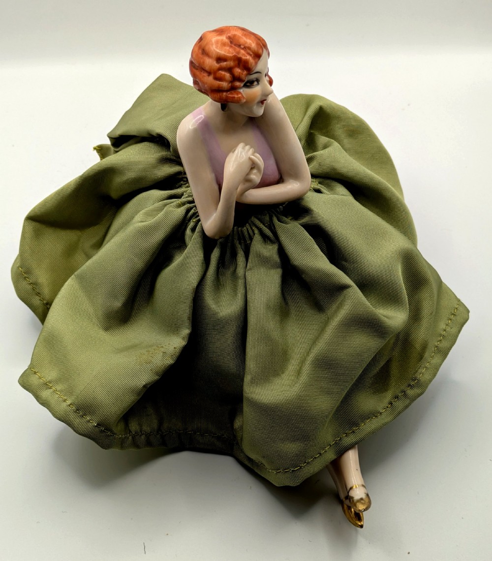 An Art Deco porcelain half doll depicting a flapper girl with auburn bobbed hair, a pink bodice, a voluminous sage green fabric skirt, and fine porcelain legs. View VIII