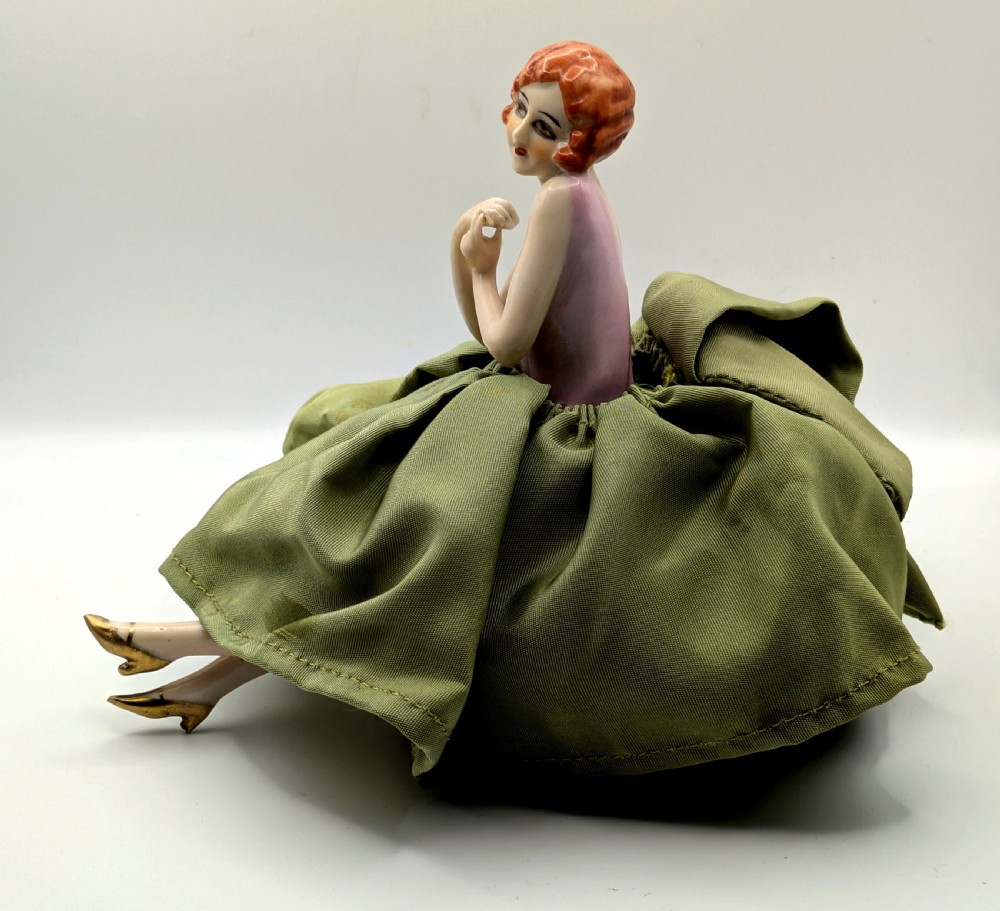 An Art Deco porcelain half doll depicting a flapper girl with auburn bobbed hair, a pink bodice, a voluminous sage green fabric skirt, and fine porcelain legs. View IX