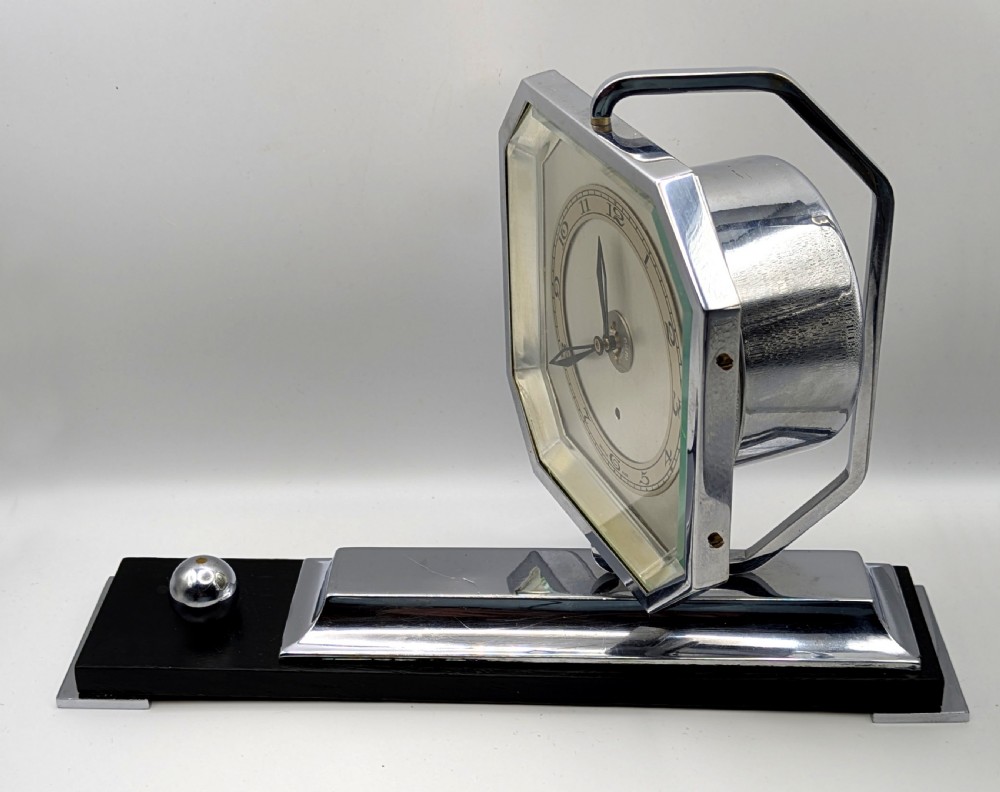 An Art Deco desk clock featuring an octagonal chrome frame mounted on a stepped base with an ebonised plinth and a small chrome sphere detail. View XII