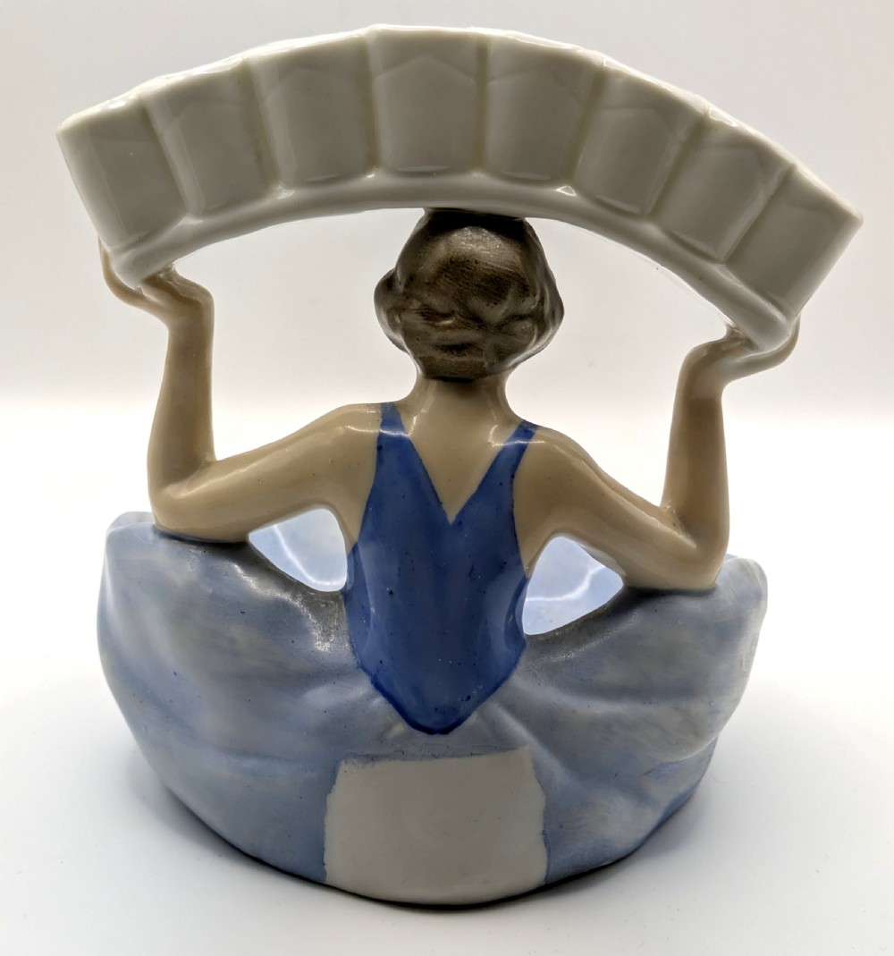An Art Deco porcelain figurine of a flapper girl wearing a blue dress and holding an arched white and red cigarette holder above her head. View III