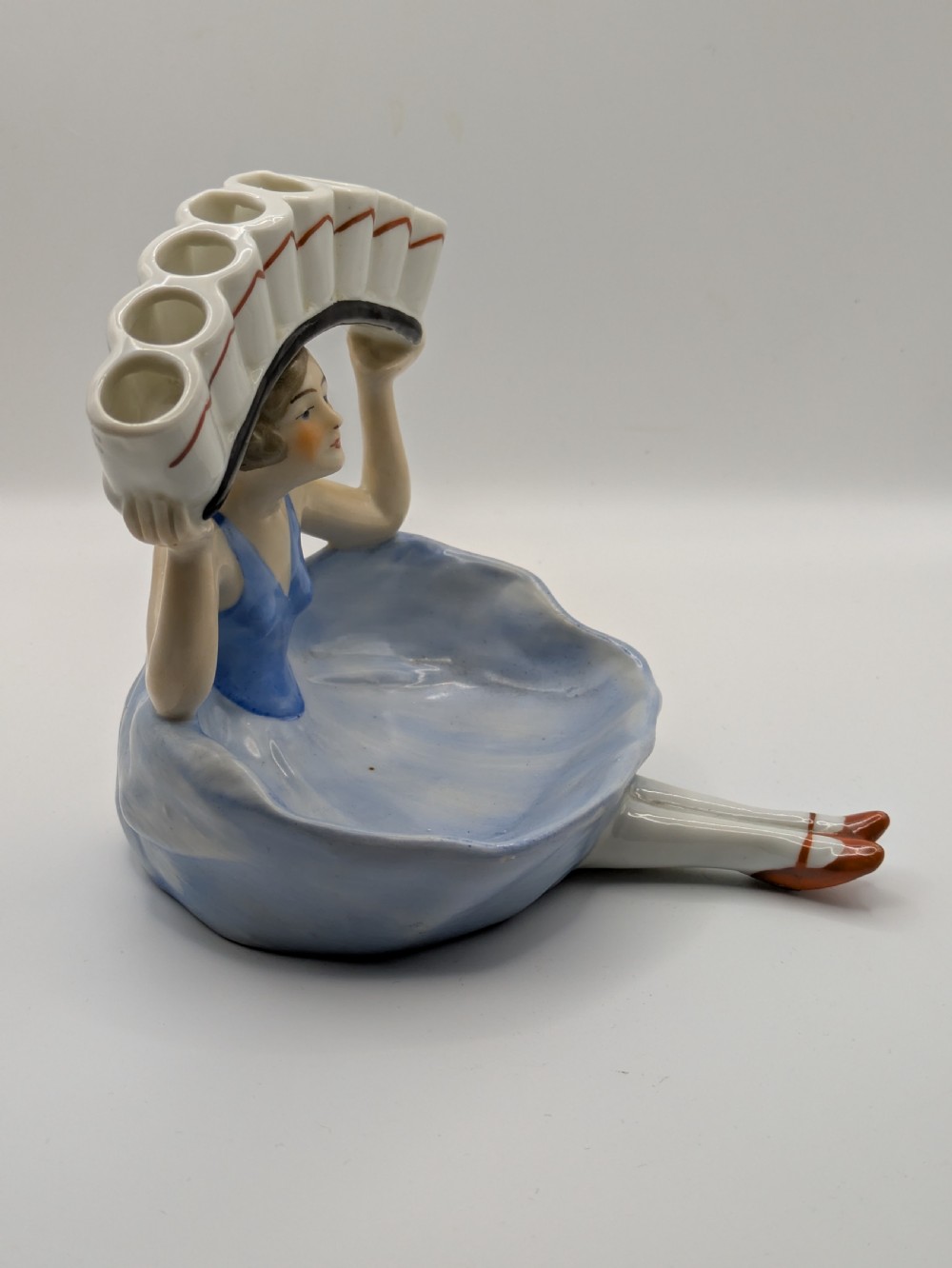 An Art Deco porcelain figurine of a flapper girl wearing a blue dress and holding an arched white and red cigarette holder above her head. View VI