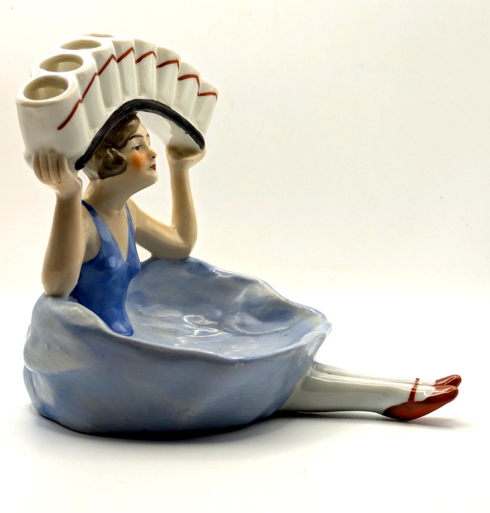An Art Deco porcelain figurine of a flapper girl wearing a blue dress and holding an arched white and red cigarette holder above her head. View VII
