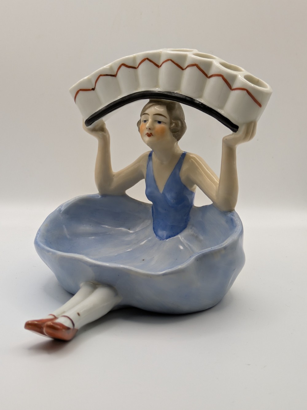 An Art Deco porcelain figurine of a flapper girl wearing a blue dress and holding an arched white and red cigarette holder above her head. View X