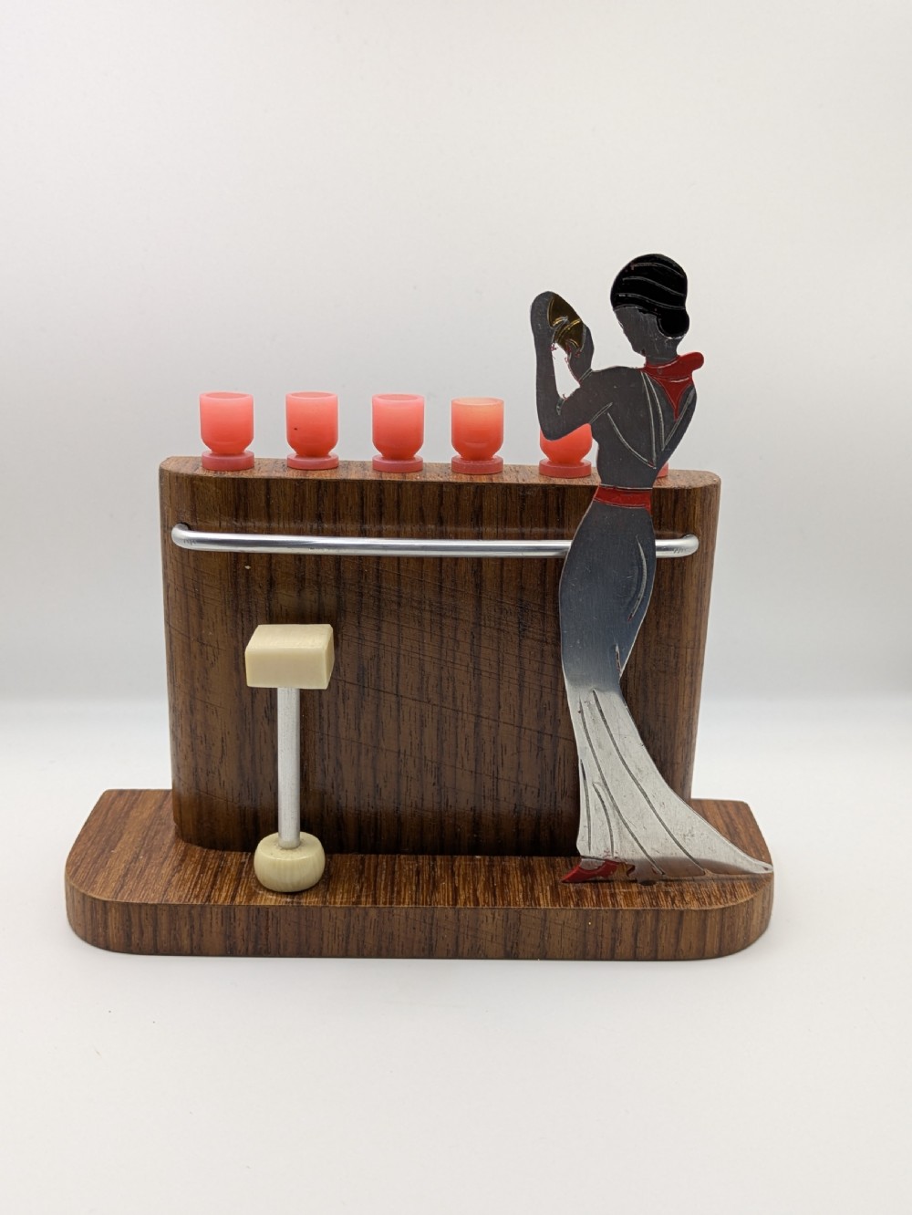 An Art Deco wooden bar stand holding red cocktail picks designed as glasses, with a cut-out silver and red female figure shaking a cocktail shaker. View IX
