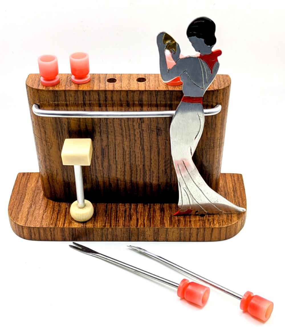 An Art Deco wooden bar stand holding red cocktail picks designed as glasses, with a cut-out silver and red female figure shaking a cocktail shaker. View X