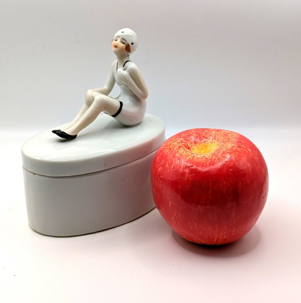 A white ceramic oval powder box featuring a sculpted Art Deco bathing belle figure wearing a white swimsuit and black shoes seated on the lid. View IX