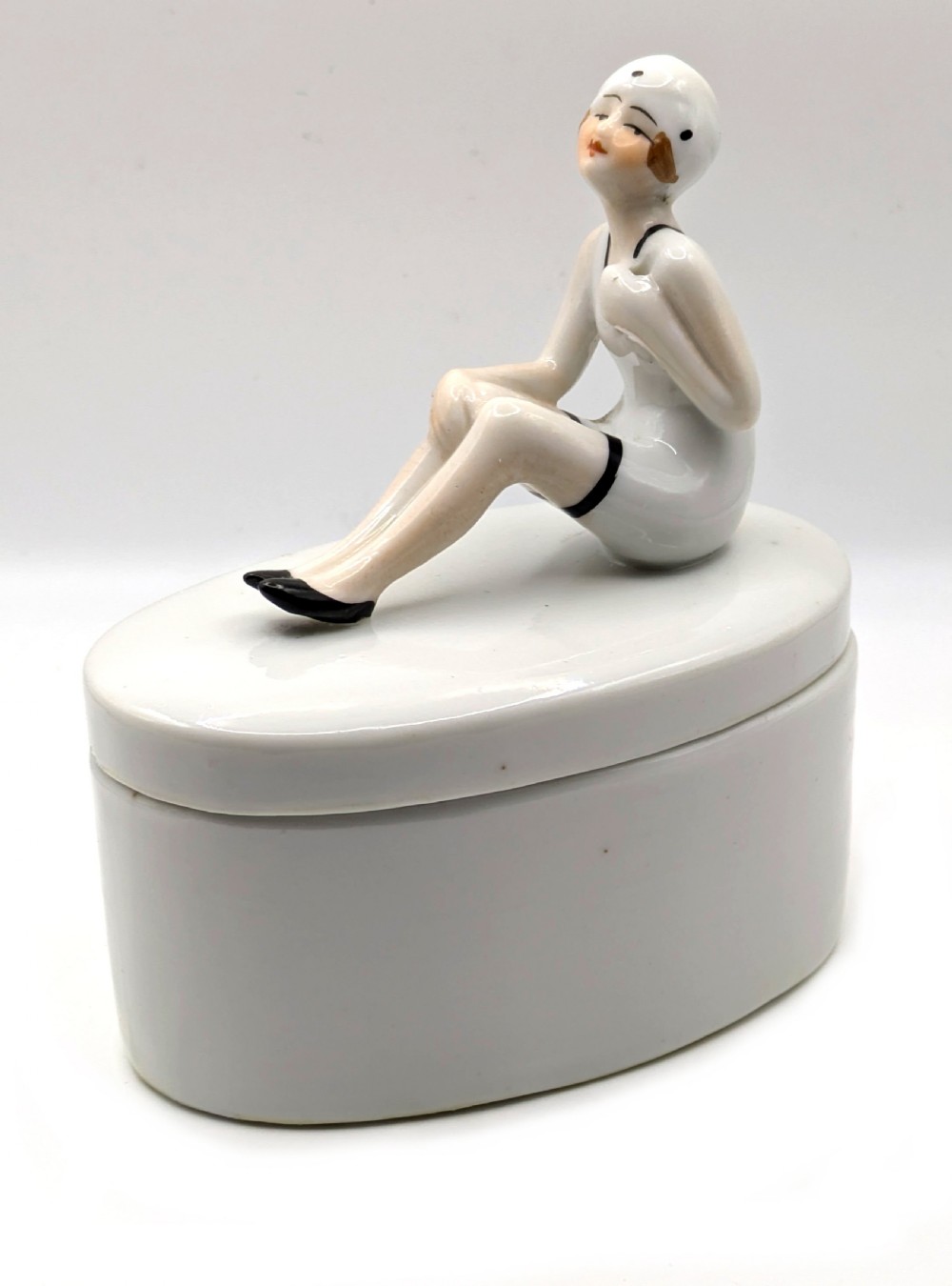 A white ceramic oval powder box featuring a sculpted Art Deco bathing belle figure wearing a white swimsuit and black shoes seated on the lid. View X