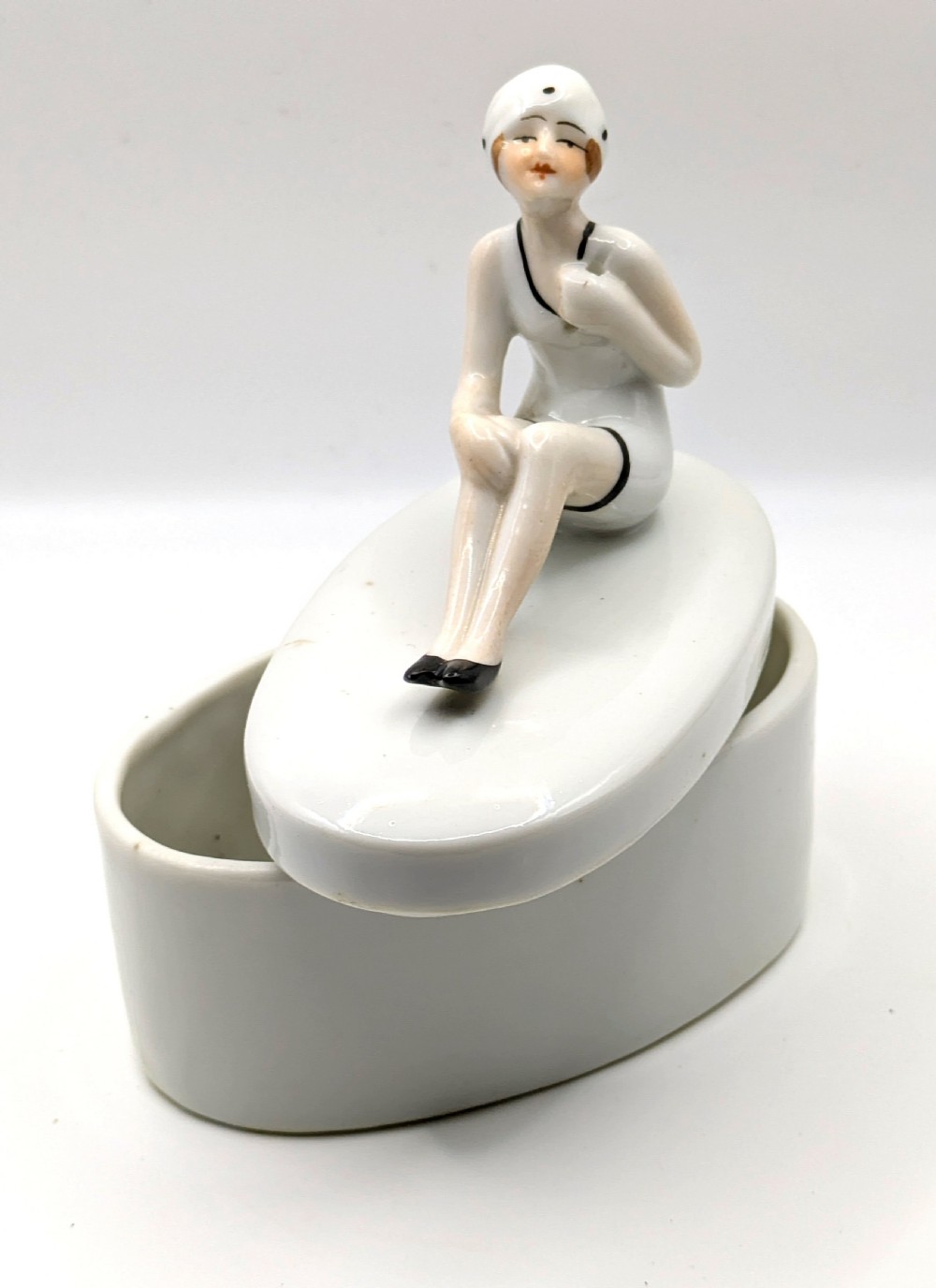 A white ceramic oval powder box featuring a sculpted Art Deco bathing belle figure wearing a white swimsuit and black shoes seated on the lid. View XI