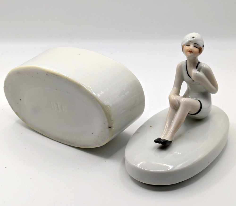 A white ceramic oval powder box featuring a sculpted Art Deco bathing belle figure wearing a white swimsuit and black shoes seated on the lid. View XII