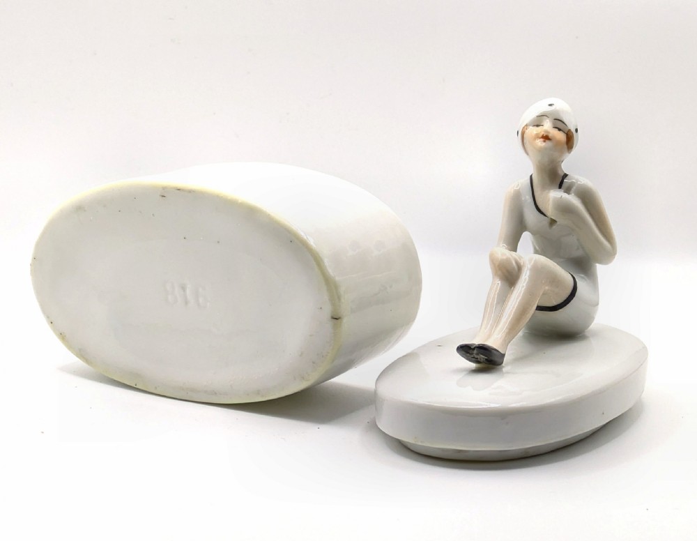 A white ceramic oval powder box featuring a sculpted Art Deco bathing belle figure wearing a white swimsuit and black shoes seated on the lid. View XIII