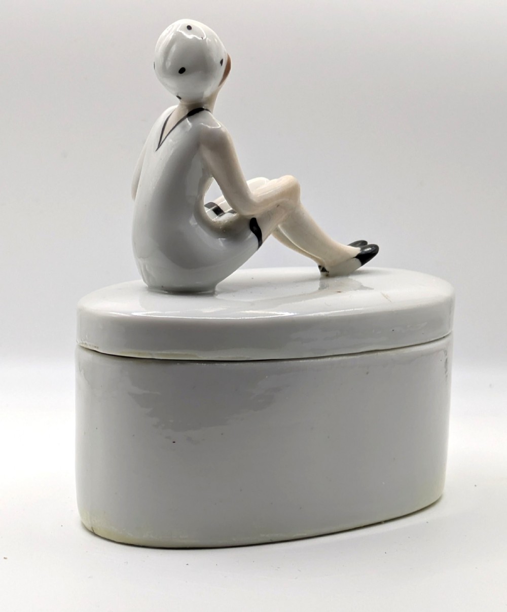 A white ceramic oval powder box featuring a sculpted Art Deco bathing belle figure wearing a white swimsuit and black shoes seated on the lid. View XIV