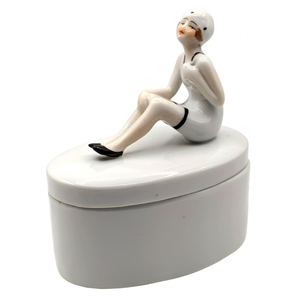 A white ceramic oval powder box featuring a sculpted Art Deco bathing belle figure wearing a white swimsuit and black shoes seated on the lid.