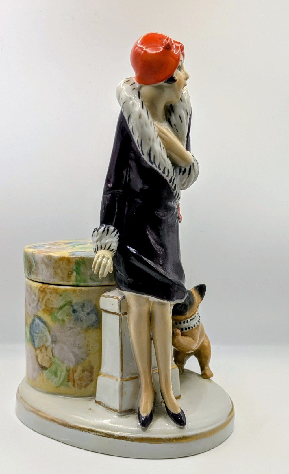 An Art Deco painted porcelain trinket box featuring a flapper lady in a black coat and red cloche hat alongside a small dog. View X