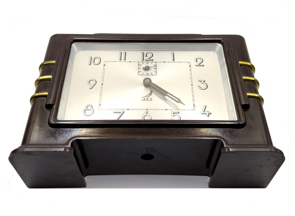 A dark brown rectangular Bakelite mantel clock with gold brass side accents and a silver rectangular dial displaying Art Deco numerals. View VII