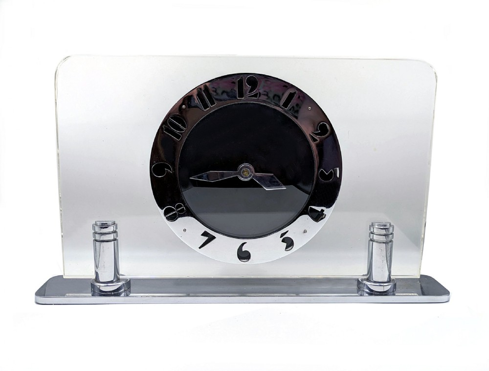 An Art Deco mantel clock featuring a clear glass rectangular panel supported by chrome columns on a metal base, with a central black dial surrounded by cutout silver numerals.