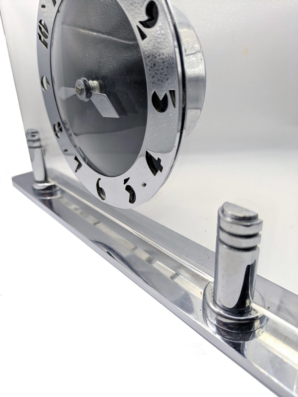 An Art Deco mantel clock featuring a clear glass rectangular panel supported by chrome columns on a metal base, with a central black dial surrounded by cutout silver numerals. View II