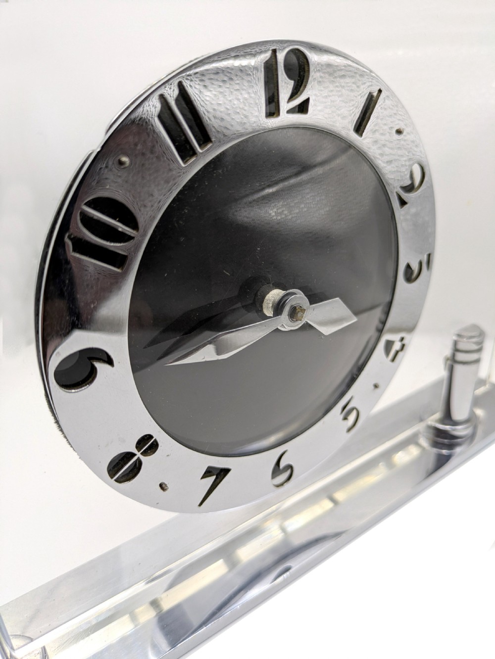 An Art Deco mantel clock featuring a clear glass rectangular panel supported by chrome columns on a metal base, with a central black dial surrounded by cutout silver numerals. View IV