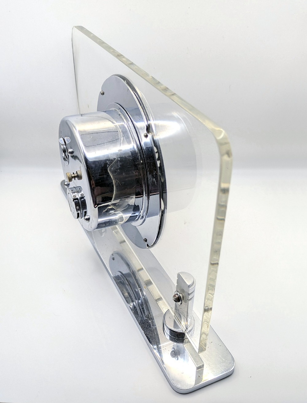 An Art Deco mantel clock featuring a clear glass rectangular panel supported by chrome columns on a metal base, with a central black dial surrounded by cutout silver numerals. View V