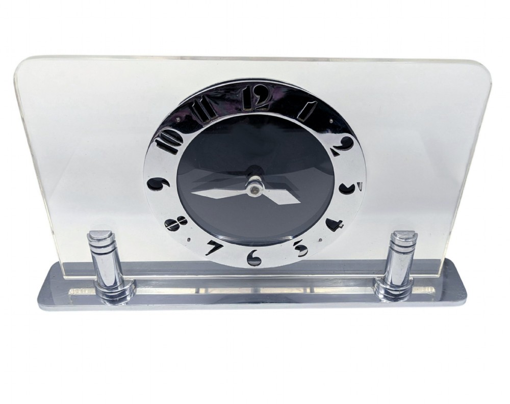 An Art Deco mantel clock featuring a clear glass rectangular panel supported by chrome columns on a metal base, with a central black dial surrounded by cutout silver numerals. View VI