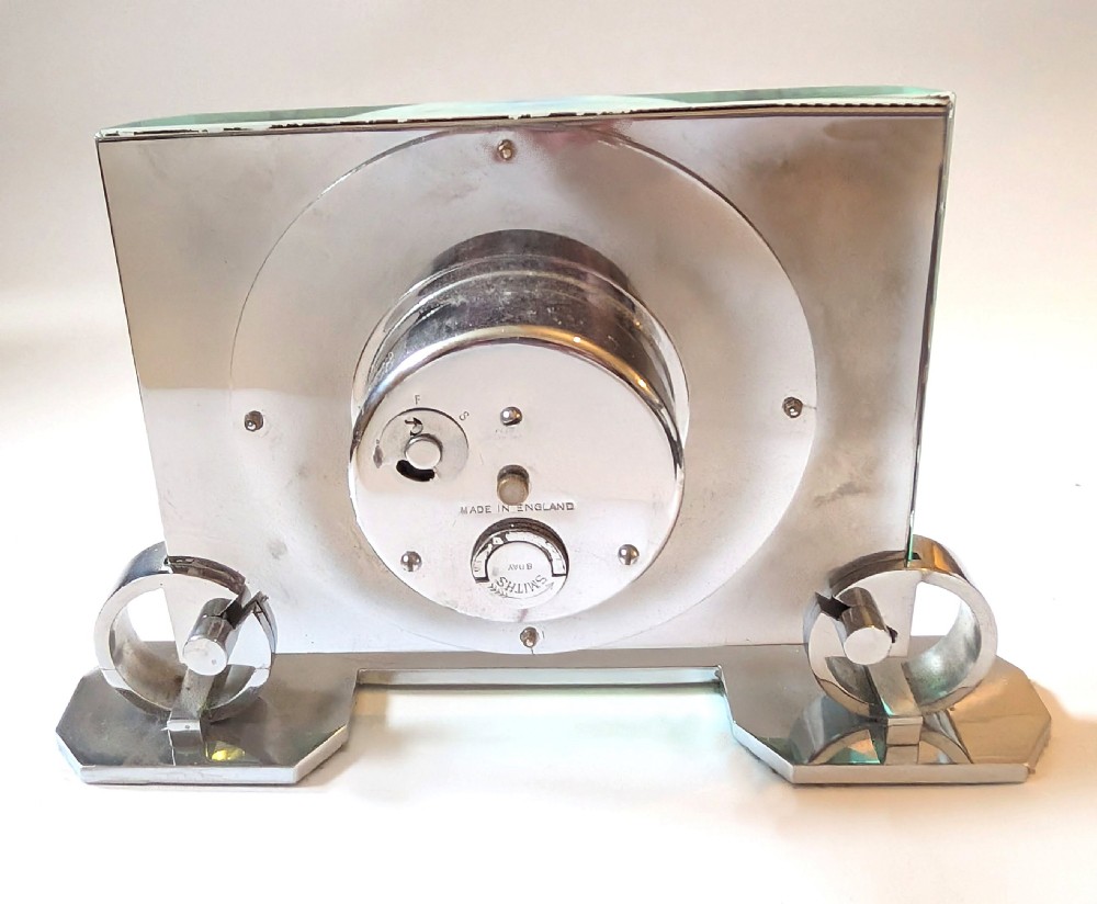 An Art Deco mantel clock featuring a thick rectangular glass body held by chrome scroll mounts on a flat metal base. View XI
