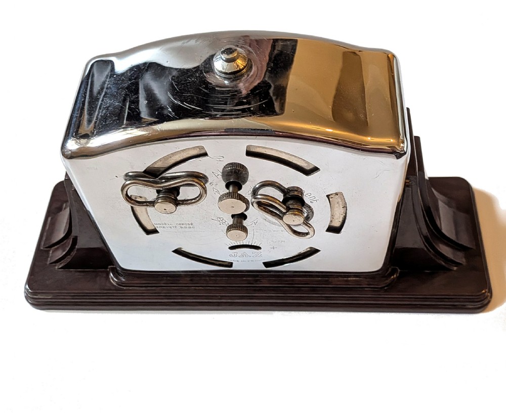 An Art Deco desk clock featuring a dark brown Bakelite stepped base, a polished chrome frame, and a cream dial with geometric Arabic numerals. View VII