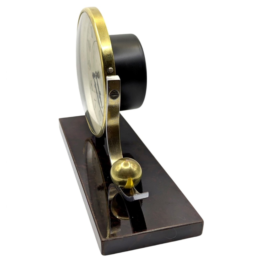 An Art Deco desk clock featuring a circular brass dial and stylised wave element resting on a rectangular dark brown Bakelite base. View IV
