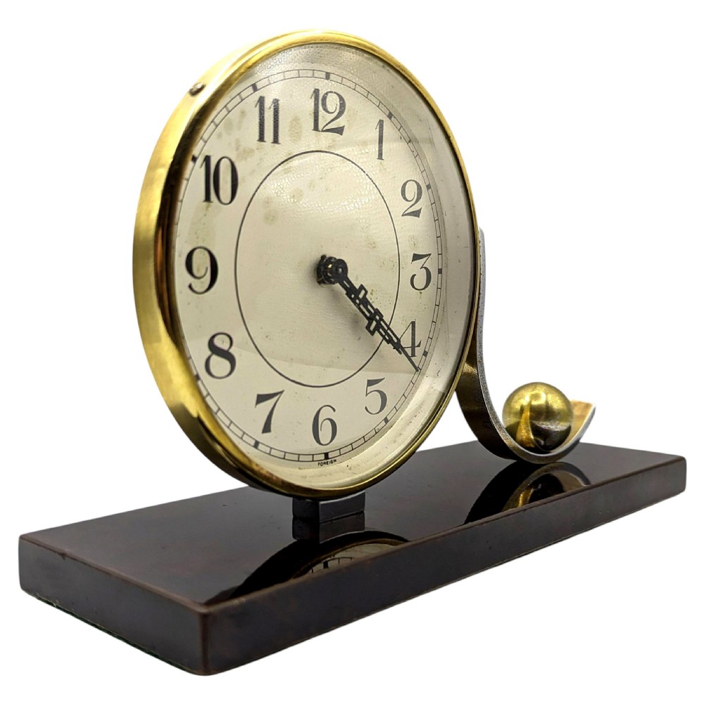 An Art Deco desk clock featuring a circular brass dial and stylised wave element resting on a rectangular dark brown Bakelite base. View V