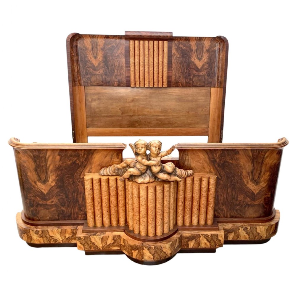 An Art Deco walnut bed featuring bookmatched veneer panels, a tall rounded headboard, and a low footboard decorated with central carved cherubs and fluted details.