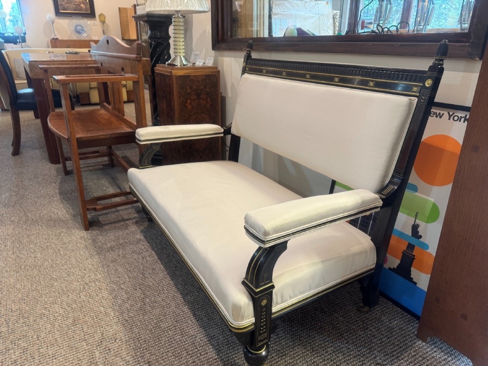 An Aesthetic Movement ebonised wooden settee featuring gilt incised line decorations and cream upholstery on the seat and backrest.