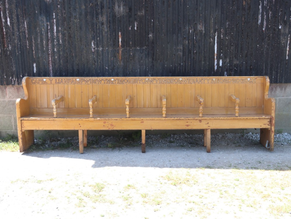 A long Victorian painted pine chapel bench featuring a vertical panelled back, turned arm dividers, and Welsh script along the top rail.
