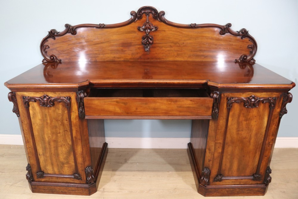 A Victorian mahogany twin pedestal sideboard featuring a central frieze drawer, carved splashback, and flame mahogany cupboard doors on plinth bases. View V
