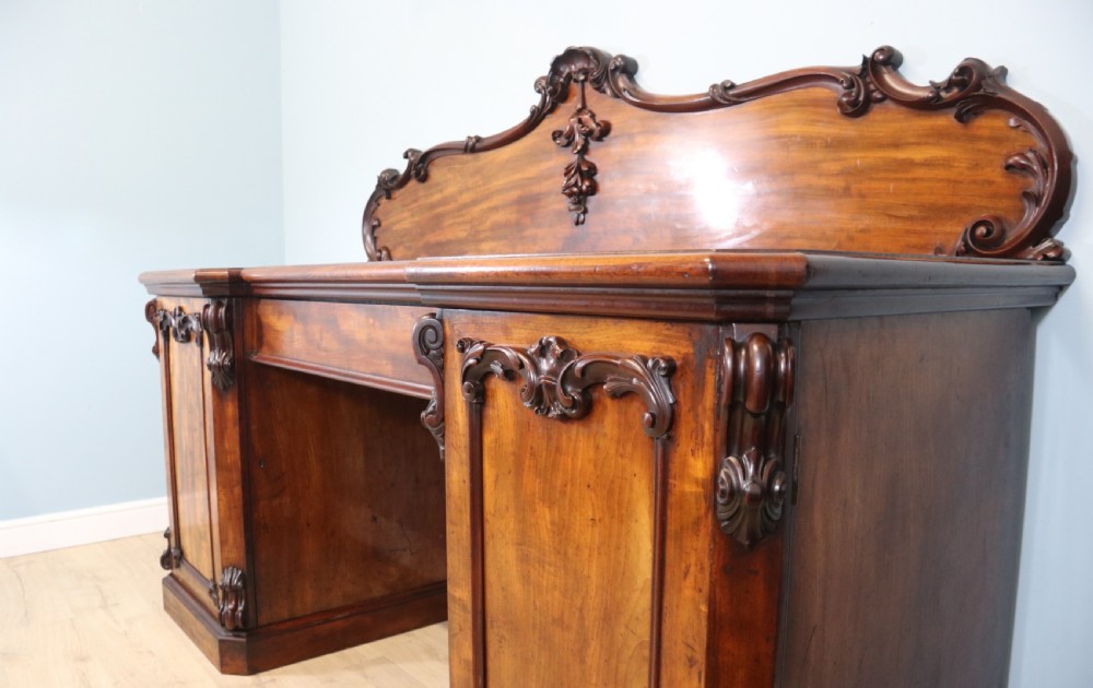 A Victorian mahogany twin pedestal sideboard featuring a central frieze drawer, carved splashback, and flame mahogany cupboard doors on plinth bases. View XVIII