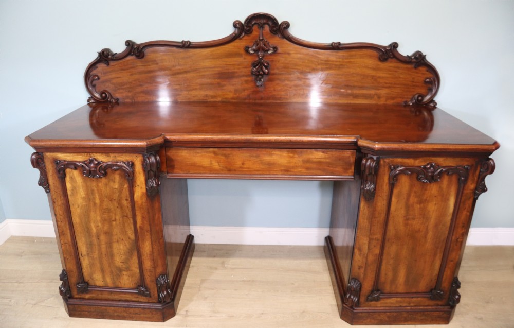 A Victorian mahogany twin pedestal sideboard featuring a central frieze drawer, carved splashback, and flame mahogany cupboard doors on plinth bases.