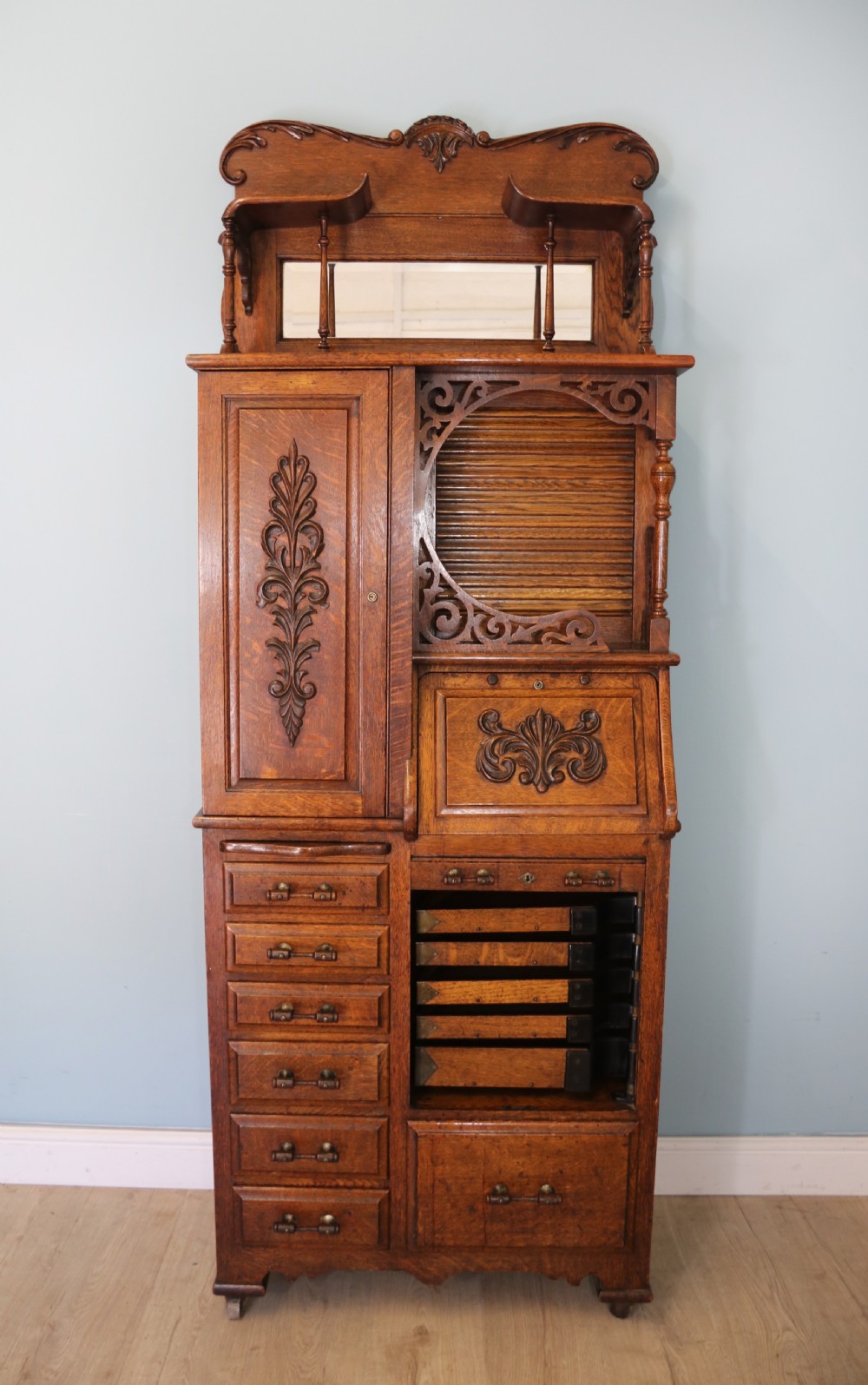 A late nineteenth-century oak combination dentist cabinet featuring a mirrored gallery top, carved foliate door, tambour section, and multiple small drawers.
