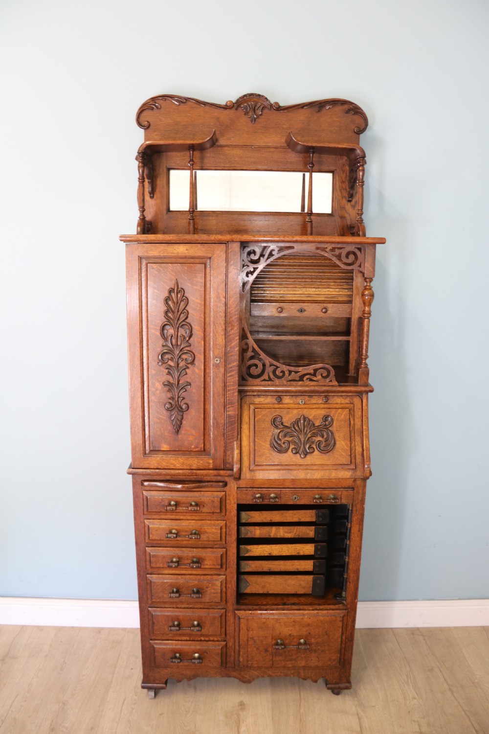 A late nineteenth-century oak combination dentist cabinet featuring a mirrored gallery top, carved foliate door, tambour section, and multiple small drawers. View II