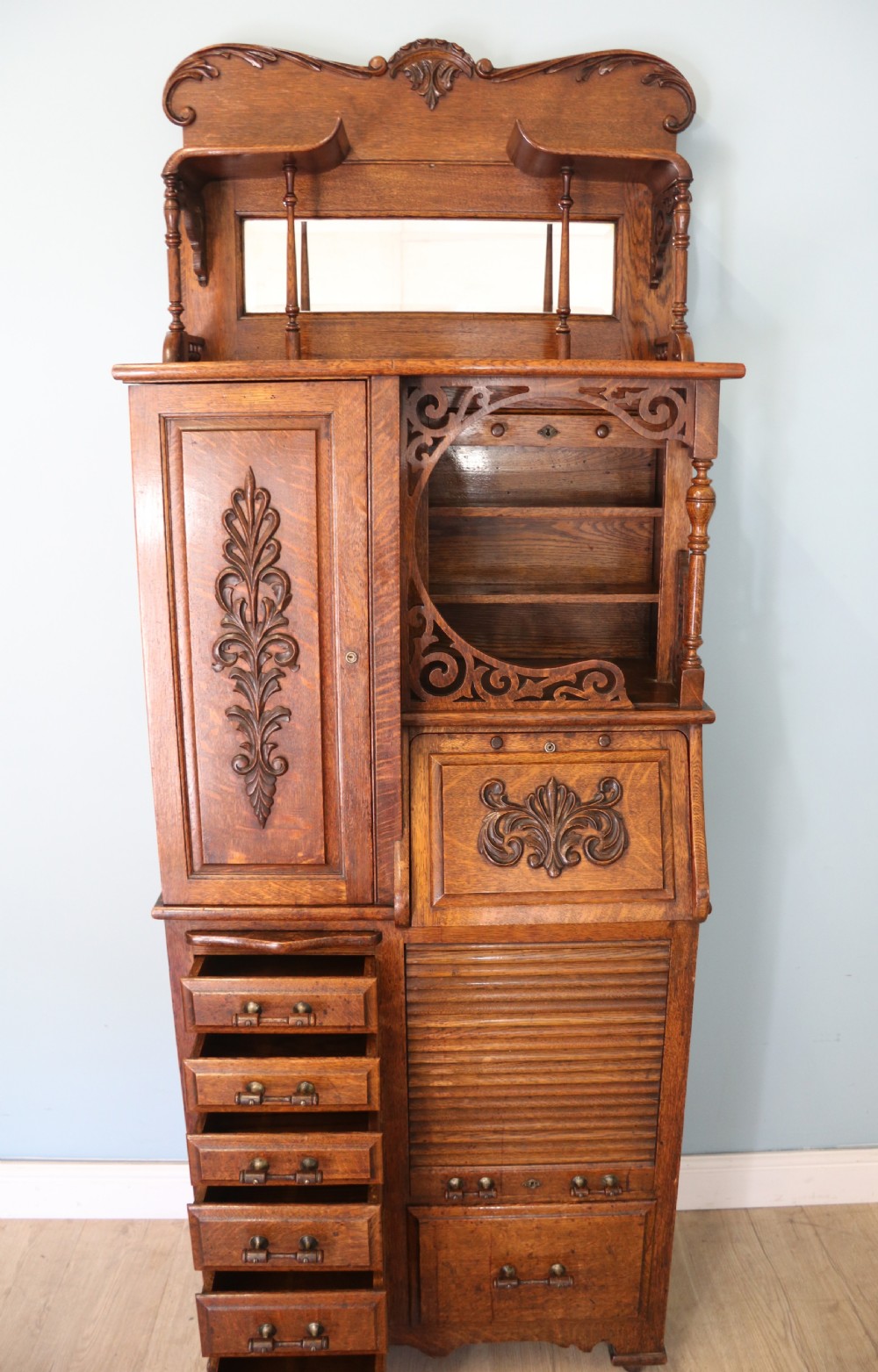 A late nineteenth-century oak combination dentist cabinet featuring a mirrored gallery top, carved foliate door, tambour section, and multiple small drawers. View IV